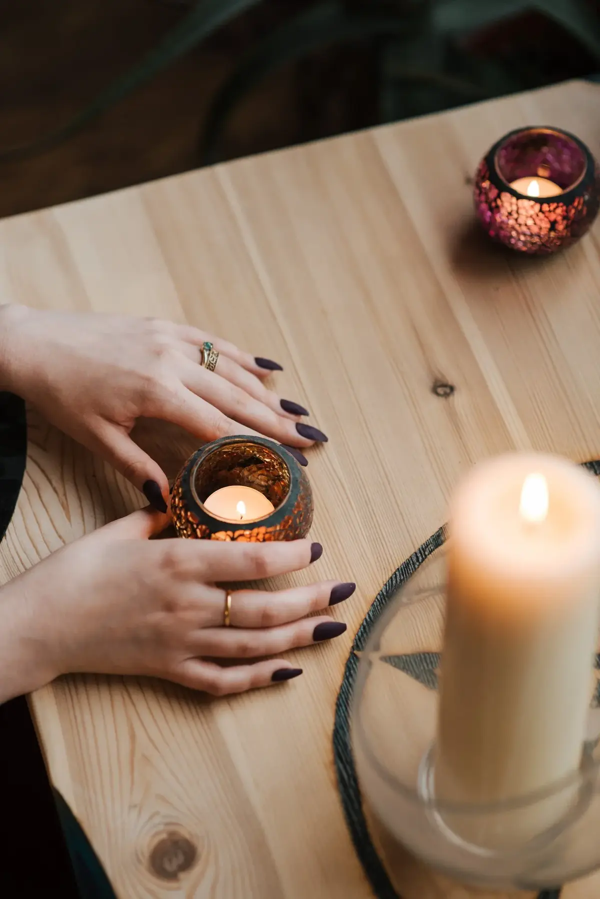 Kobiece dłonie z czarnymi paznokciami trzymające świeczkę na blacie stołu, na którym są ułożone świeczki