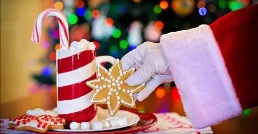 Ręka Mikołaja trzymająca świąteczny pierniczek, obok znajduje się czerwono-biała świeczka, a w tle jest choinka