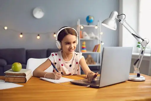 Dziewczynka siedzi przed komputerem ze słuchawkami na uszach, robiąc notatki w zeszycie